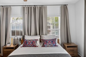 Une chambre à coucher comprenant un lit, deux tables de chevet, une commode, une fenêtre avec des stores et des rideaux.