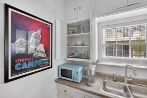 A kitchen with a framed advertisement, a blue microwave, a coffee maker, and a window with a view. - Museum & Med District Retreat 2BR + Parking (Houston)