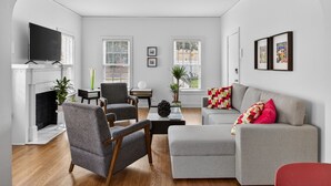 A living room with a fireplace, a television, a grey sofa, a red chair, and a coffee table. - Museum & Med District Retreat 2BR + Parking (Houston)