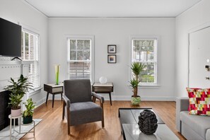 Un salon avec un fauteuil gris, une table basse, un téléviseur et deux fenêtres équipées de stores.