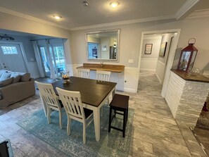 Ein Essbereich mit Holztisch und weißen Stühlen, eine Küche mit weiß gefliester Wand und ein Wohnzimmer mit beigefarbenem Sofa.