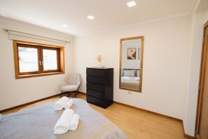 Een slaapkamer met een bed, een stoel, een dressoir, een raam en een spiegel.