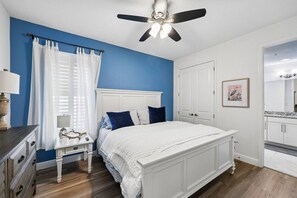 Ein Schlafzimmer mit einem Bett, einem Nachttisch, einem Deckenventilator, einem Fenster mit Vorhängen und einer Tür.