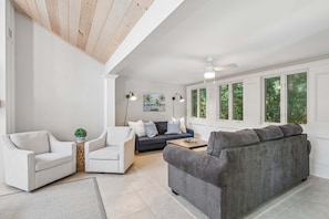 Een ruime woonkamer met een grijze bank, witte fauteuils en een donkere bank. Er staan twee vloerlampen, een plafondventilator en een groot raam met uitzicht op bomen.