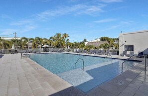 A rectangular pool with a metal ladder and railings, surrounded by lounge chairs and umbrellas, with palm trees and a building in the background. - Exec Bay Resort-Style 4BR Townhome w/Beach, Marina, Heat/Chill Pool & Pickleball (Islamorada)