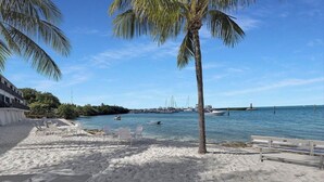 A beach with white sand, palm trees, and a view of boats and a pier in the distance. - Exec Bay Resort-Style 4BR Townhome w/Beach, Marina, Heat/Chill Pool & Pickleball (Islamorada)