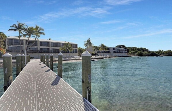 A pier leading to a building with a beach view, palm trees, and clear water. - Exec Bay Resort-Style 4BR Townhome w/Beach, Marina, Heat/Chill Pool & Pickleball (Islamorada)