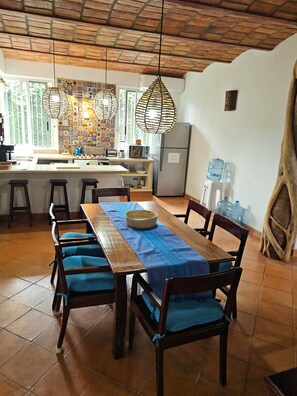 Ein Essbereich mit einem Holztisch, blauen gepolsterten Stühlen, einer Hängelampe und einer Küche im Hintergrund.