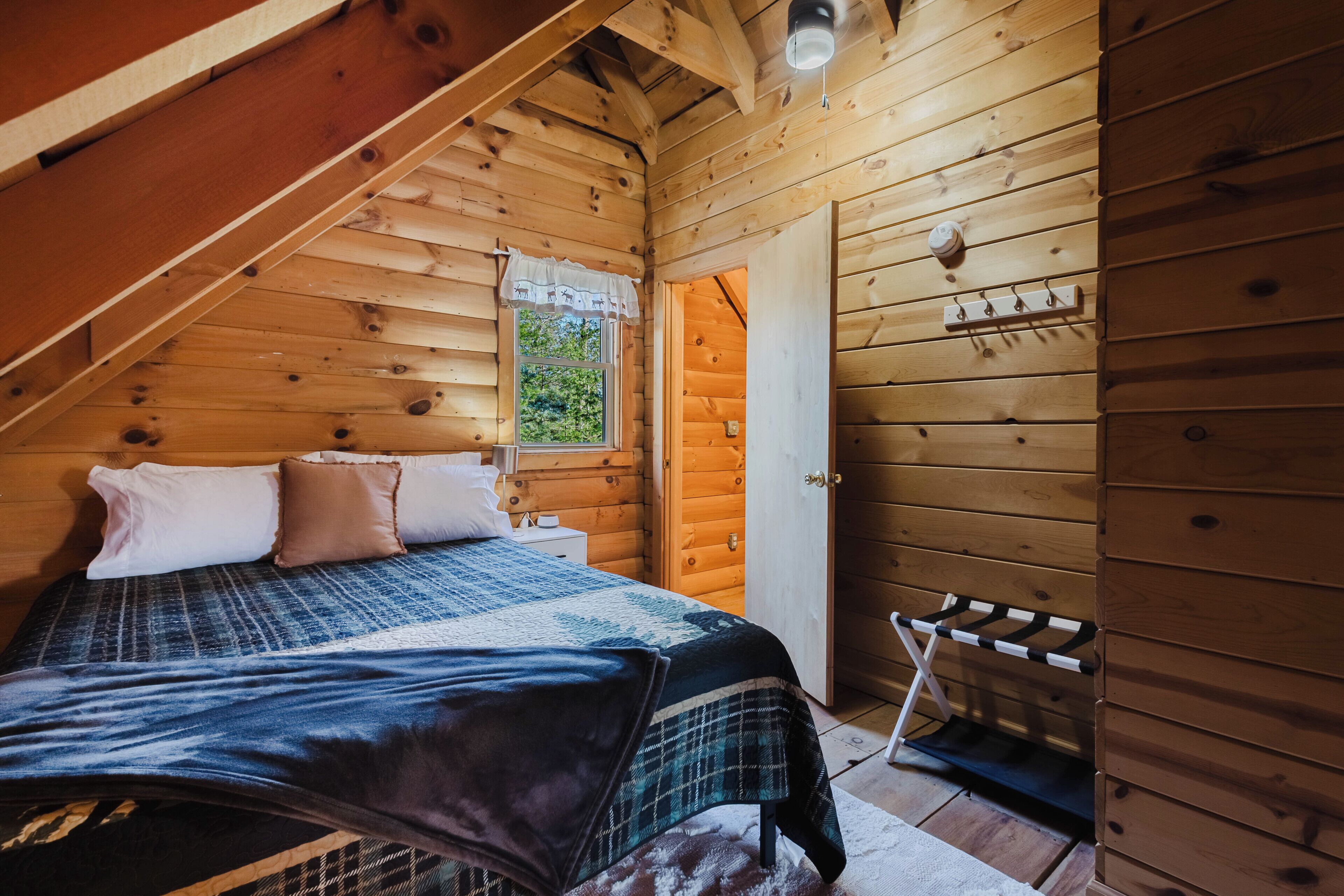 A wooden cabin bedroom with a bed, pillows, a window, and a coat rack.