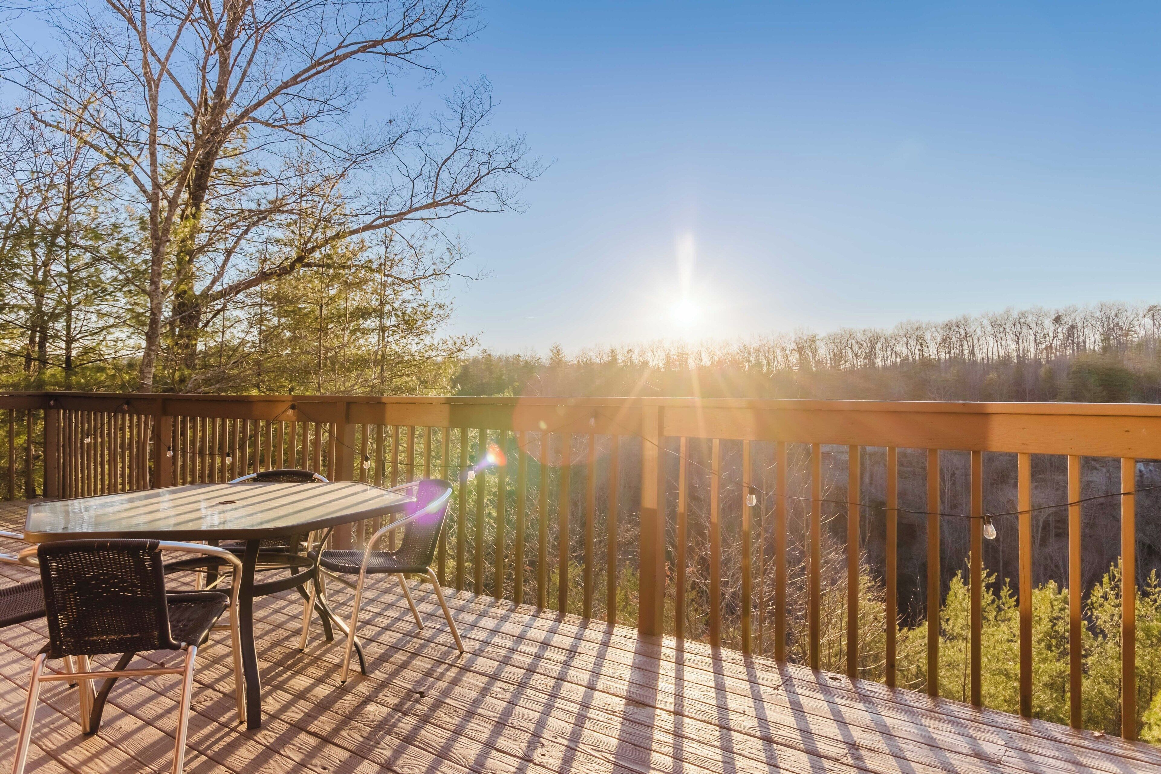 A wooden deck with a glass table and chairs, overlooking a forested area.