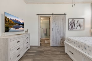 A bedroom with a wooden dresser, a bed with a floral bedspread, a sliding barn door, a framed picture of horses, and a large landscape painting on the wall.