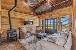 A log cabin interior with a fireplace, leather chairs, a sofa, and a large window.