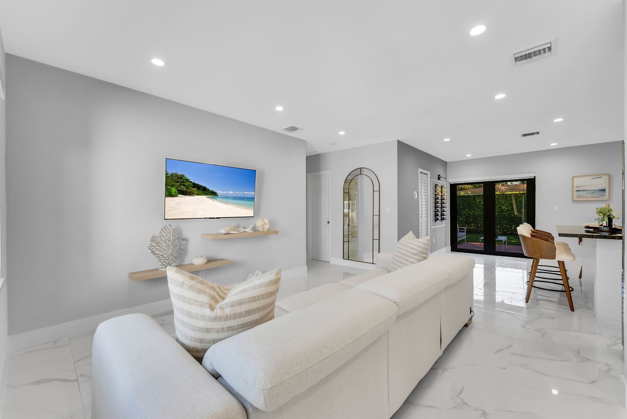 A modern living room with a large flat-screen TV, a sofa with cushions, a dining area with a chair, and a view of a garden through a glass door.
