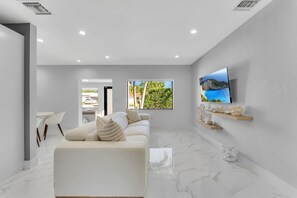 Ein modernes Wohnzimmer mit einem weißen Sofa, einem an der Wand montierten Flachbildfernseher und Blick auf eine Küstenlandschaft durch ein Fenster.