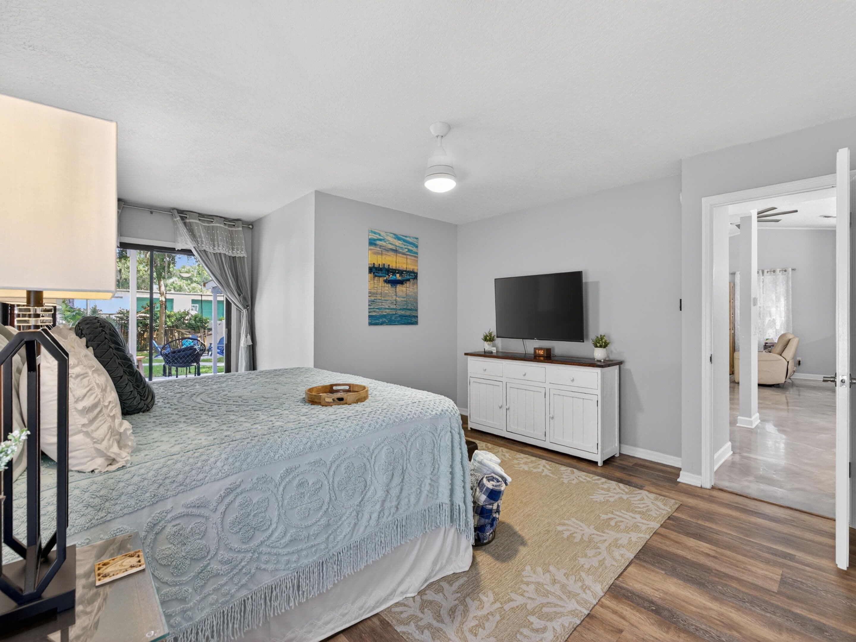 A bedroom with a bed, a TV, a painting, and a view of the outdoors.