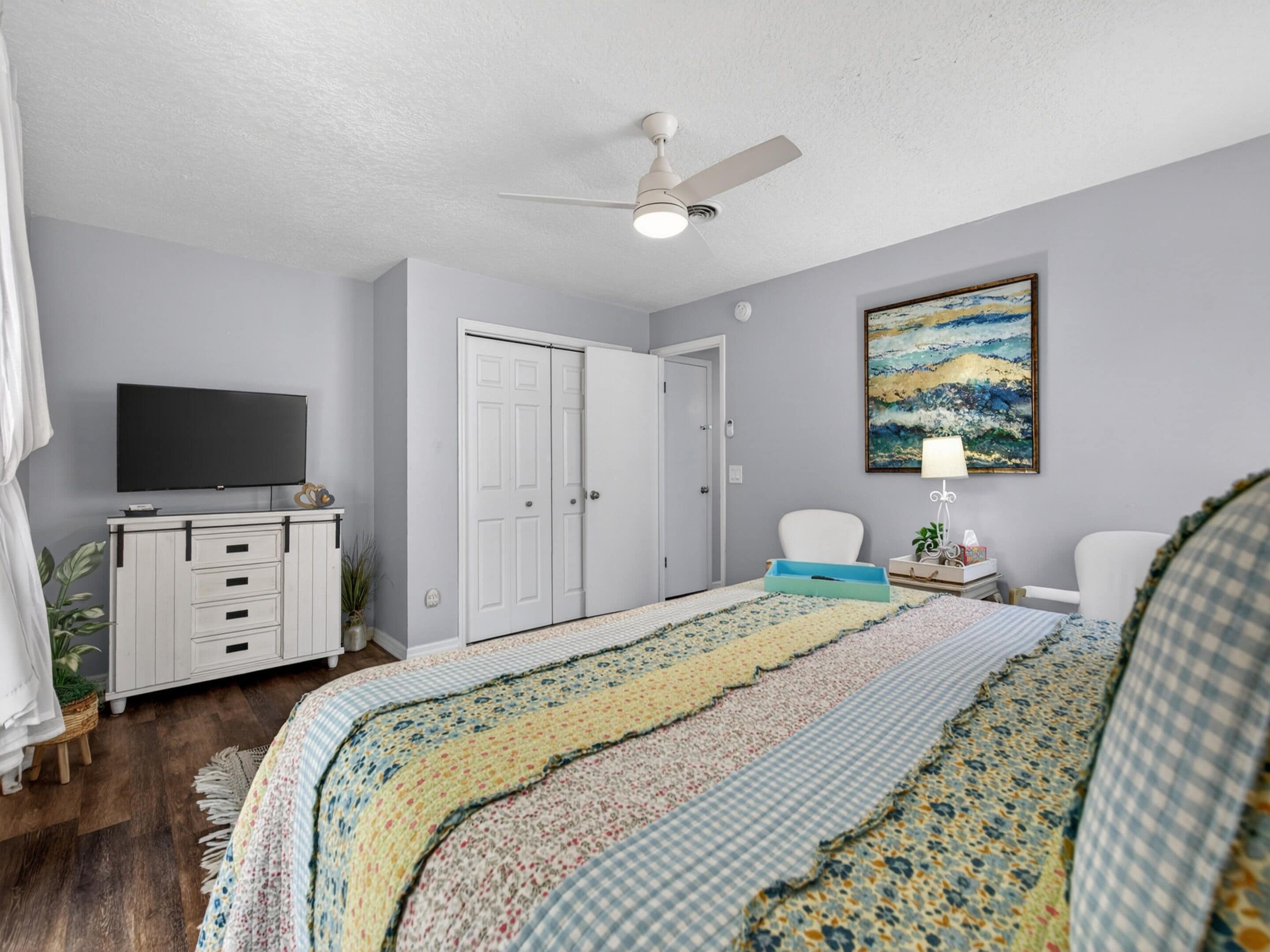 A bedroom with a bed, a television, a ceiling fan, a door, and a painting on the wall.