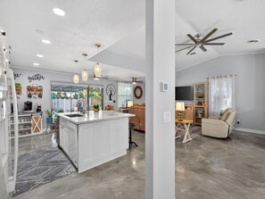 Eine moderne Küche mit Kochinsel, ein Essbereich mit Sofa und Tisch sowie ein Wohnzimmer mit Deckenventilator.