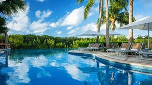 Une piscine d’un bleu éclatant, bordée de chaises longues et d’ombrelles, et entourée de palmiers et de verdure.