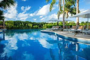 A clear blue swimming pool with lounge chairs and umbrellas, surrounded by palm trees and greenery.