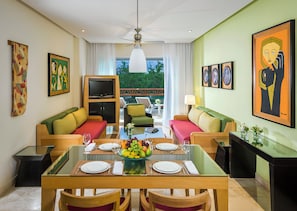 A modern living room with a dining area, featuring a glass-top table, colorful sofas, and artwork on the walls.