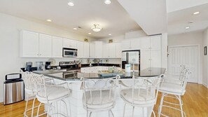 A modern kitchen with white cabinetry, a central island with bar stools, and built-in appliances. - Spacious home with large kitchen, outdoor shower, kayak launch access (Bethany Beach)