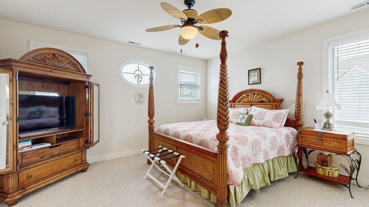 A bedroom with a four-poster bed, a television cabinet, a bedside table with a lamp, and a ceiling fan.