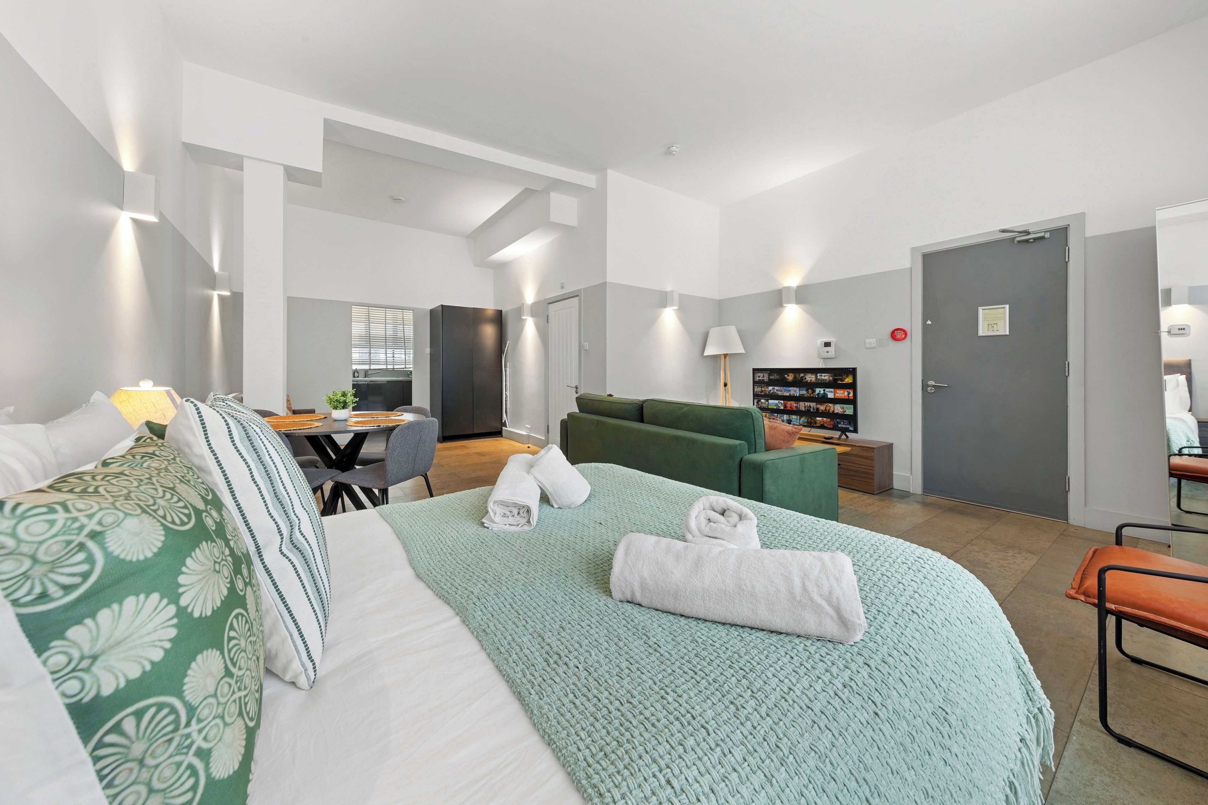 A hotel room with a bed, a sofa, a dining table with chairs, a TV, and a small kitchenette.