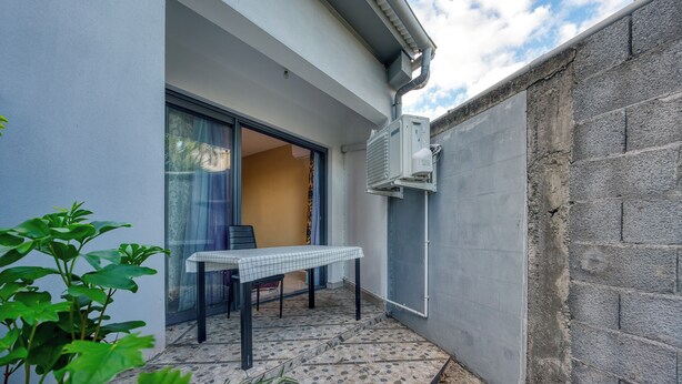 A small outdoor patio with a table and chairs, a sliding glass door leading to an interior space, and an air conditioning unit mounted on the wall. - Vétyvers - Climatisation - Terrasse (Saint-Louis)