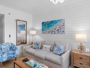 Ein Wohnzimmer mit Sofa, Sessel, Couchtisch und einem Gemälde einer Strandlandschaft.