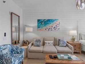 A living room with a beige sofa, a patterned armchair, a coffee table with remote controls, and a beach-themed painting on the wall. - Bahia 4122 (Miramar Beach)