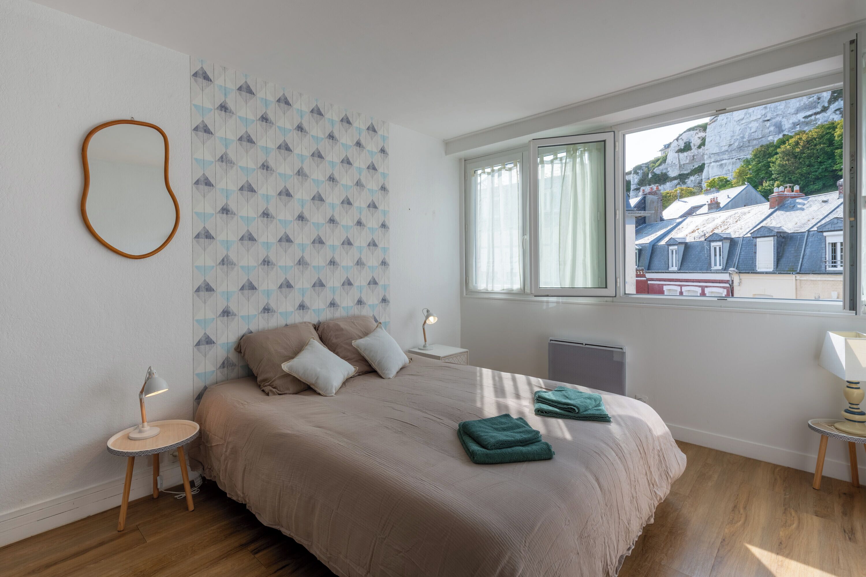 Un dormitorio con cama, mesita de noche, lámpara, ventana con vistas a edificios y pared con diseño.