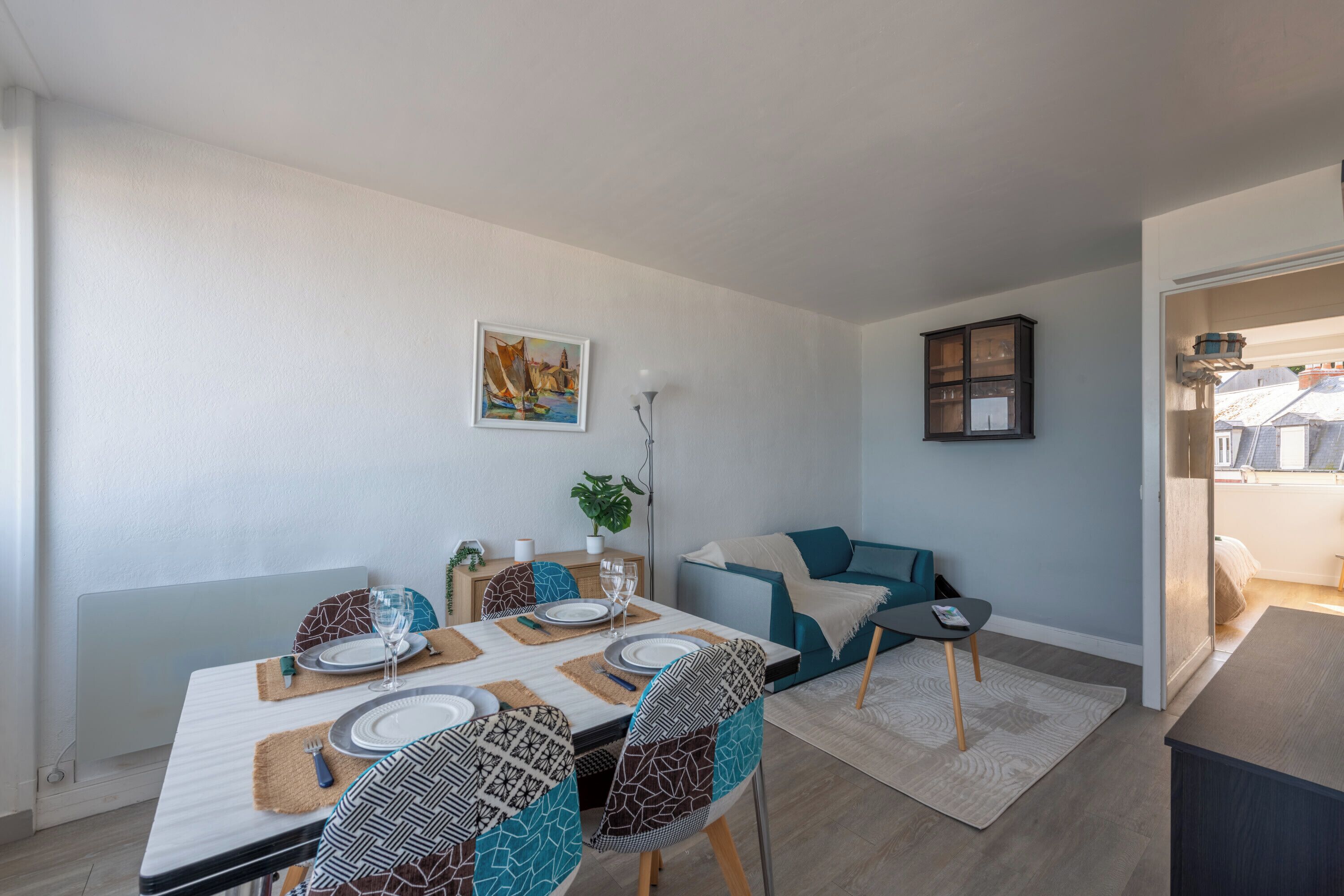 Un comedor con una mesa preparada para cuatro personas, un sofá, un armario con puertas de vidrio y un cuadro en la pared.