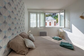 A bedroom with a large bed, a radiator, a window with curtains, and a view of buildings outside. - Le Relai de la Marée -Plage -Petfriendly (Le Tréport)