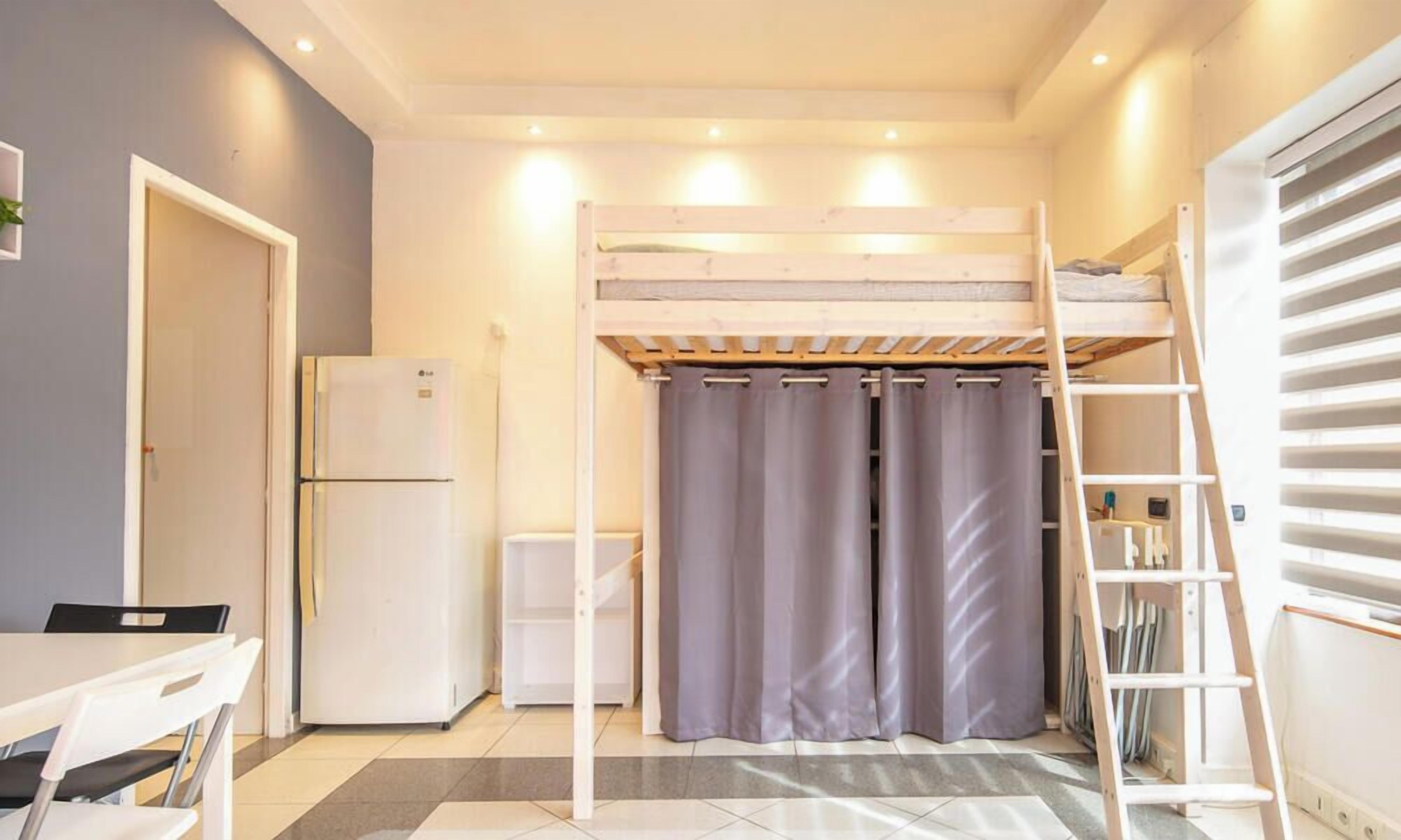 Een kleine kamer met een stapelbed, een tafeltje, een koelkast en een gordijn.