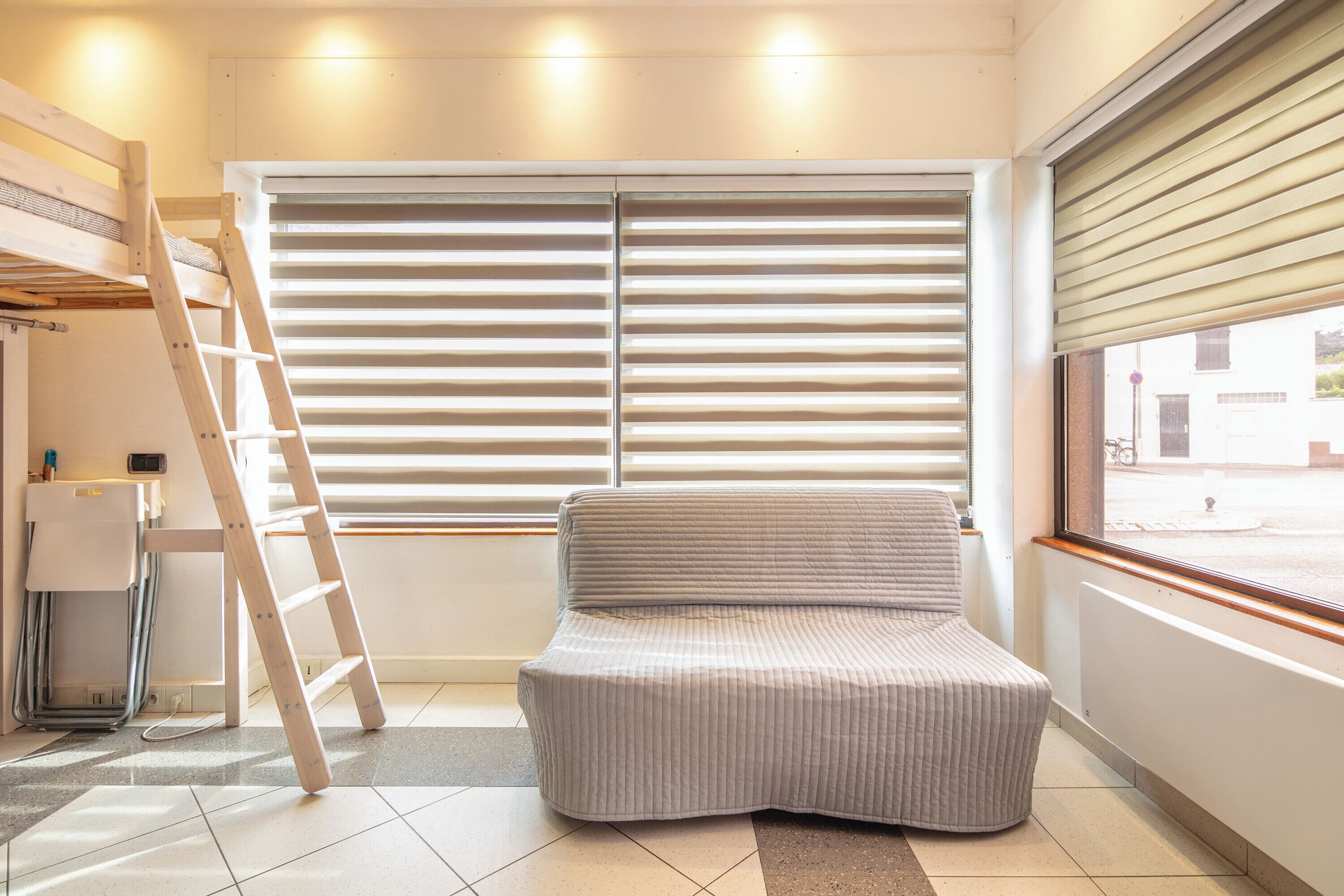Een kamer met een bed, een ladder, een raam met zonwering en uitzicht op de straat.