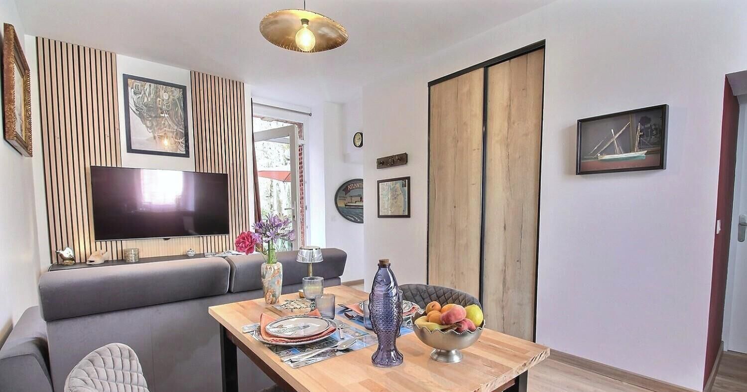 A modern living room with a flat-screen TV, a dining table with a bowl of fruit, and a grey sofa.