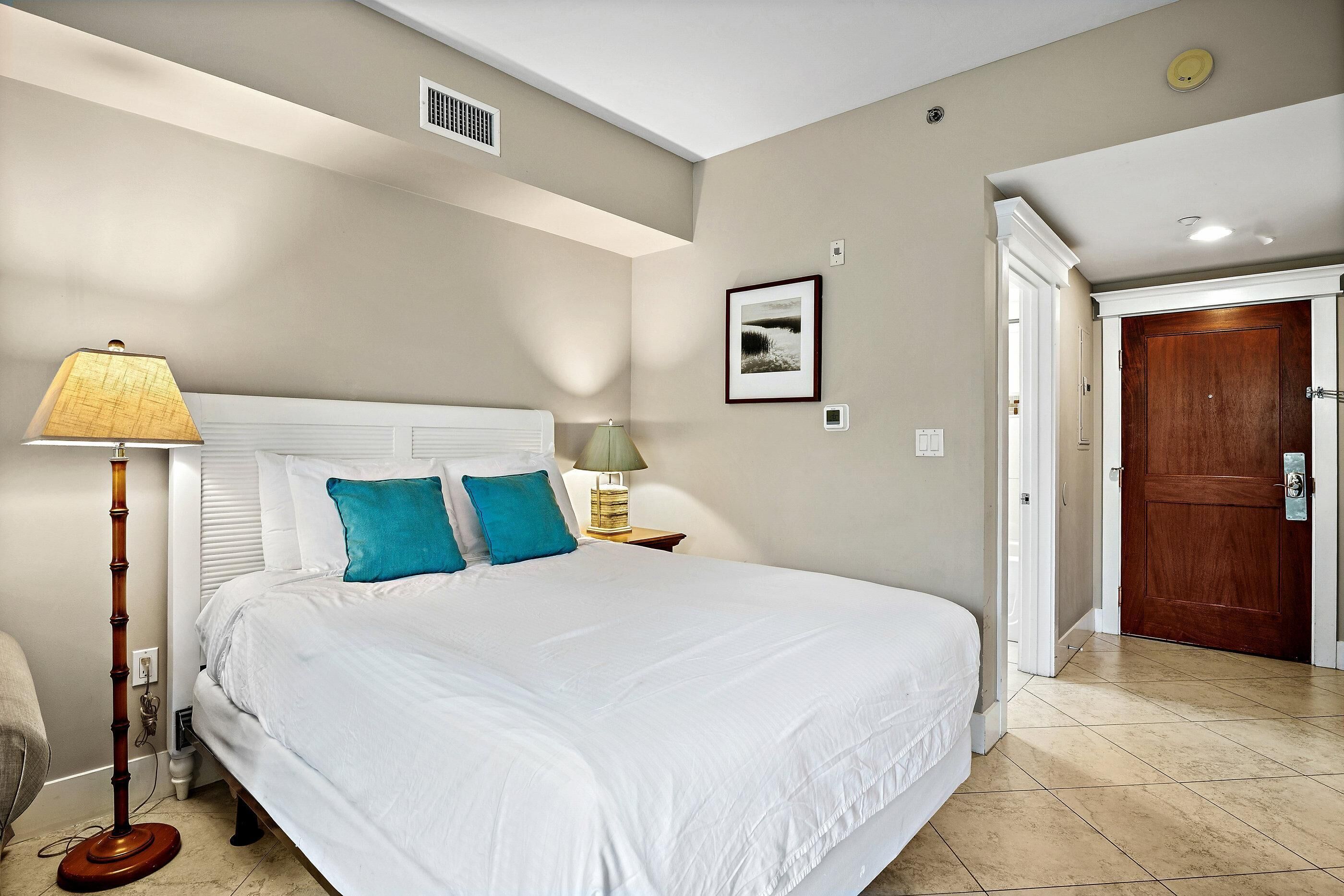 A bedroom with a large bed, a bedside lamp, a picture on the wall, and a door to the right.