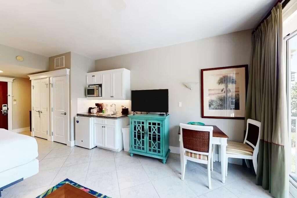 Une chambre d’hôtel comprenant un lit, une kitchenette, un téléviseur, un coin repas avec une table et des chaises, ainsi qu’une fenêtre avec des rideaux.