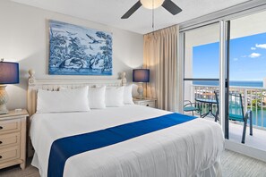 Ein Hotelzimmer mit einem großen Bett, zwei Nachttischen, einem Deckenventilator, einem Gemälde, Meerblick und einem Balkon mit Tisch und Stühlen.