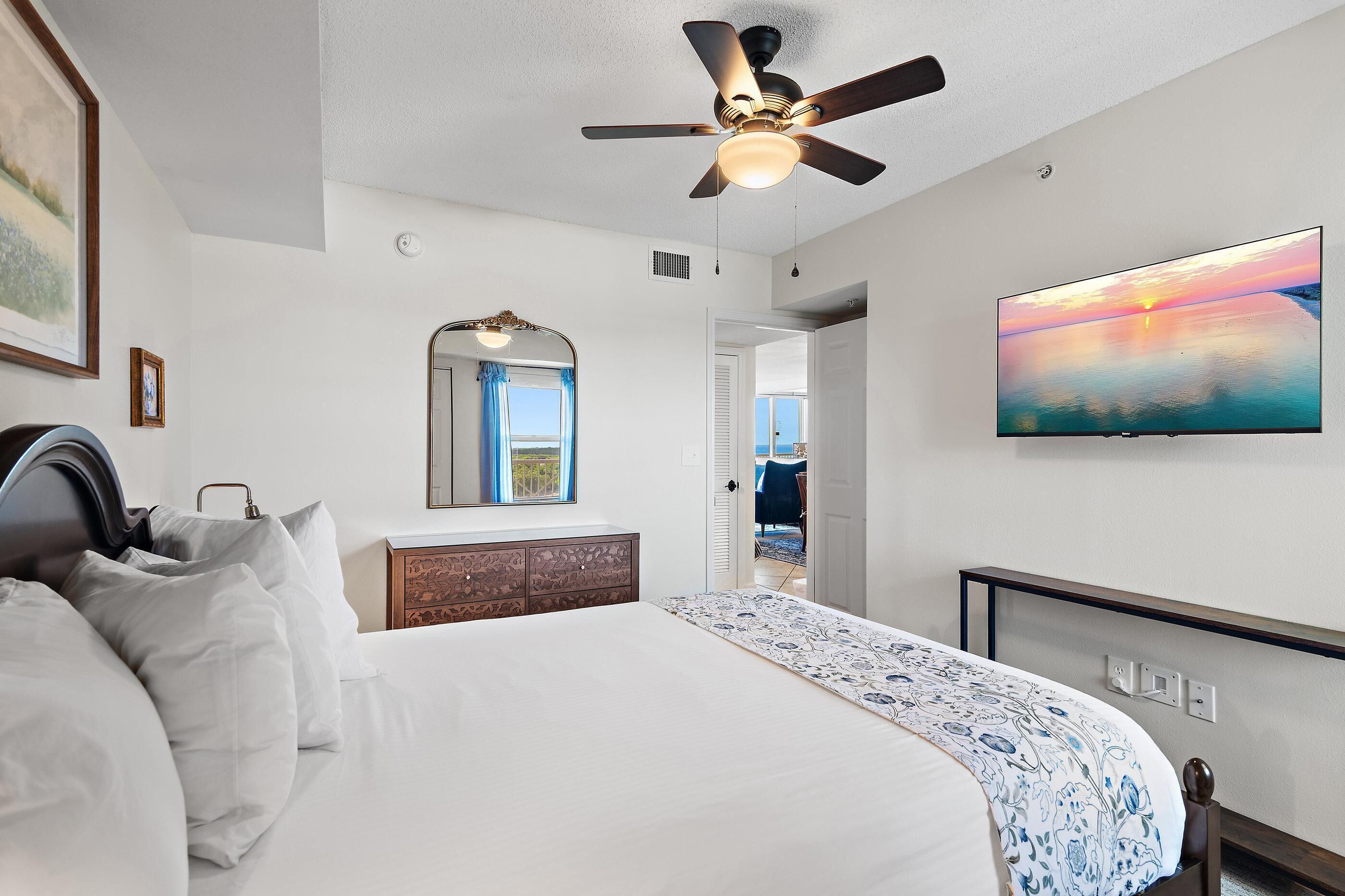 A hotel room with a bed, a ceiling fan, a large painting, a mirror, and a view of the outside.