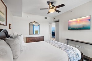 A hotel room with a bed, a ceiling fan, a large painting, a mirror, and a view of the outside. - Pelican Beach Terrace 903 (Destin)