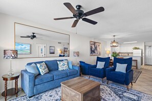 Ein Wohnzimmer mit einem blauen Sofa, zwei blauen Sesseln, einem Holztisch, einem Deckenventilator und einem großen Spiegel.