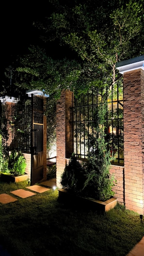 A gated entrance with brick pillars and metal bars, surrounded by lush greenery and a well-lit pathway. - Chalé Jardim Itália (Tiangua)