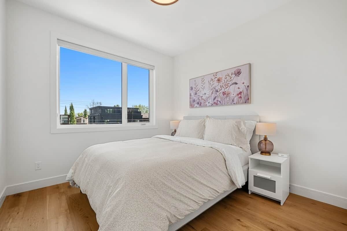 A bedroom with a large window, a bed with white bedding, a nightstand with a lamp, and a floral artwork on the wall.