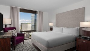 Quarto de hotel com uma cama grande, duas mesas de cabeceira com abajures, uma poltrona roxa e vista para edifícios da cidade através de uma janela ampla.