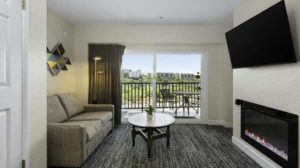 A hotel room with a sofa, a small table, a fireplace, and a balcony with a view of buildings.