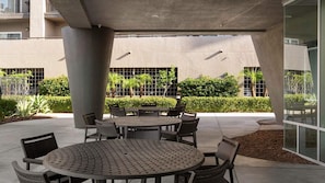 En udendørs patio med runde borde og stole, omgivet af grønt og en bygningsfacade.