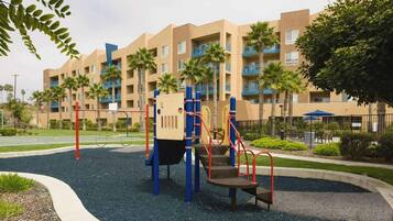Un parque infantil con estructuras para escalar y un tobogán, rodeado de un área ajardinada con palmeras y un edificio con balcones al fondo.