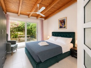 A bedroom with a bed, a desk, and a chair. There is a window with curtains and a view of greenery outside.