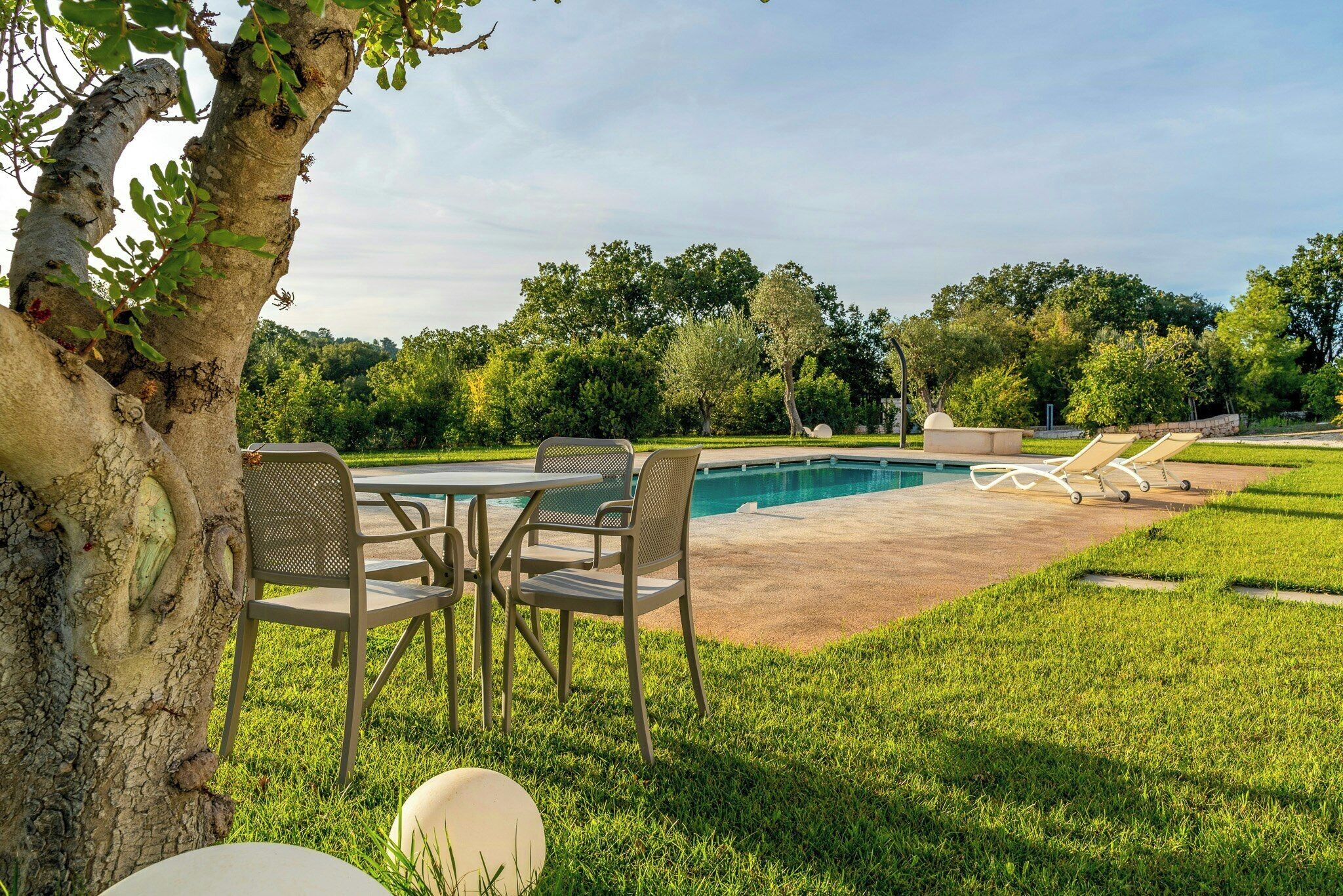A pool area with a table and chairs, surrounded by grass and trees.